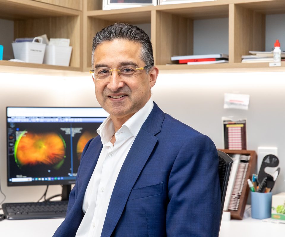 An Eastwood Eye Surgery ophthalmologist at his consulting-room desk, a wide-field retinal scan on the monitor beside him