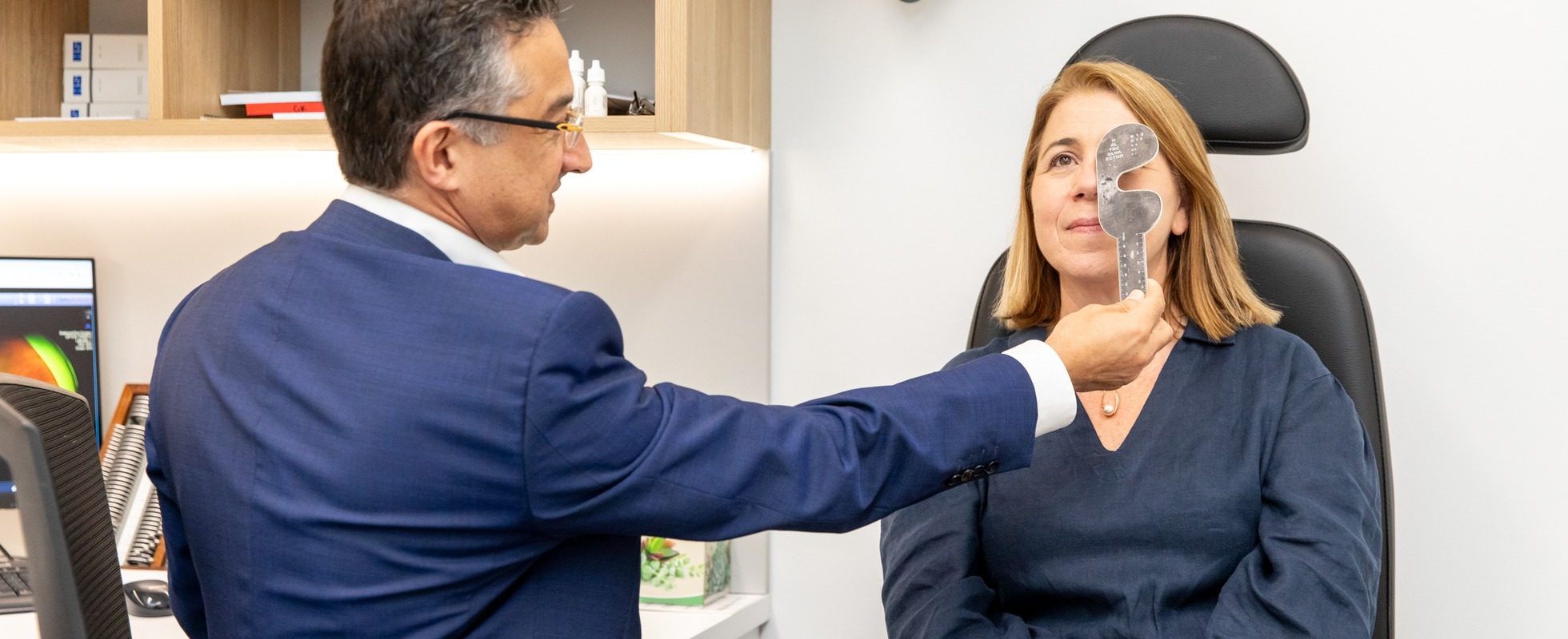 Eastwood Eye Surgery ophthalmologist checking a patient's vision with an occluder at a post-operative review, a retinal image on the monitor behind